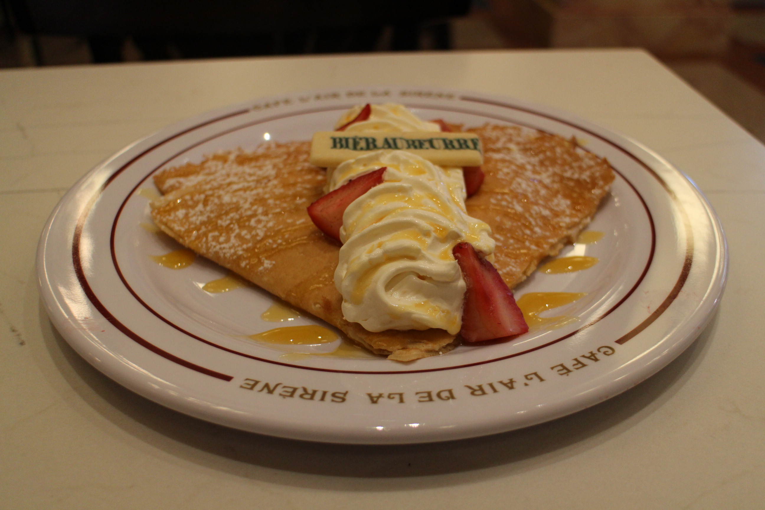 The Bieraubeurre Crepe on a white plate at Cafe L'air de la Sirène | Ministry of Magic at Epic Universe. This treat from the WIzarding World has a sweet, Butterbeer whipped topping, strawberries and a shortbread cookie with Bieraubeurre printed on it on top.