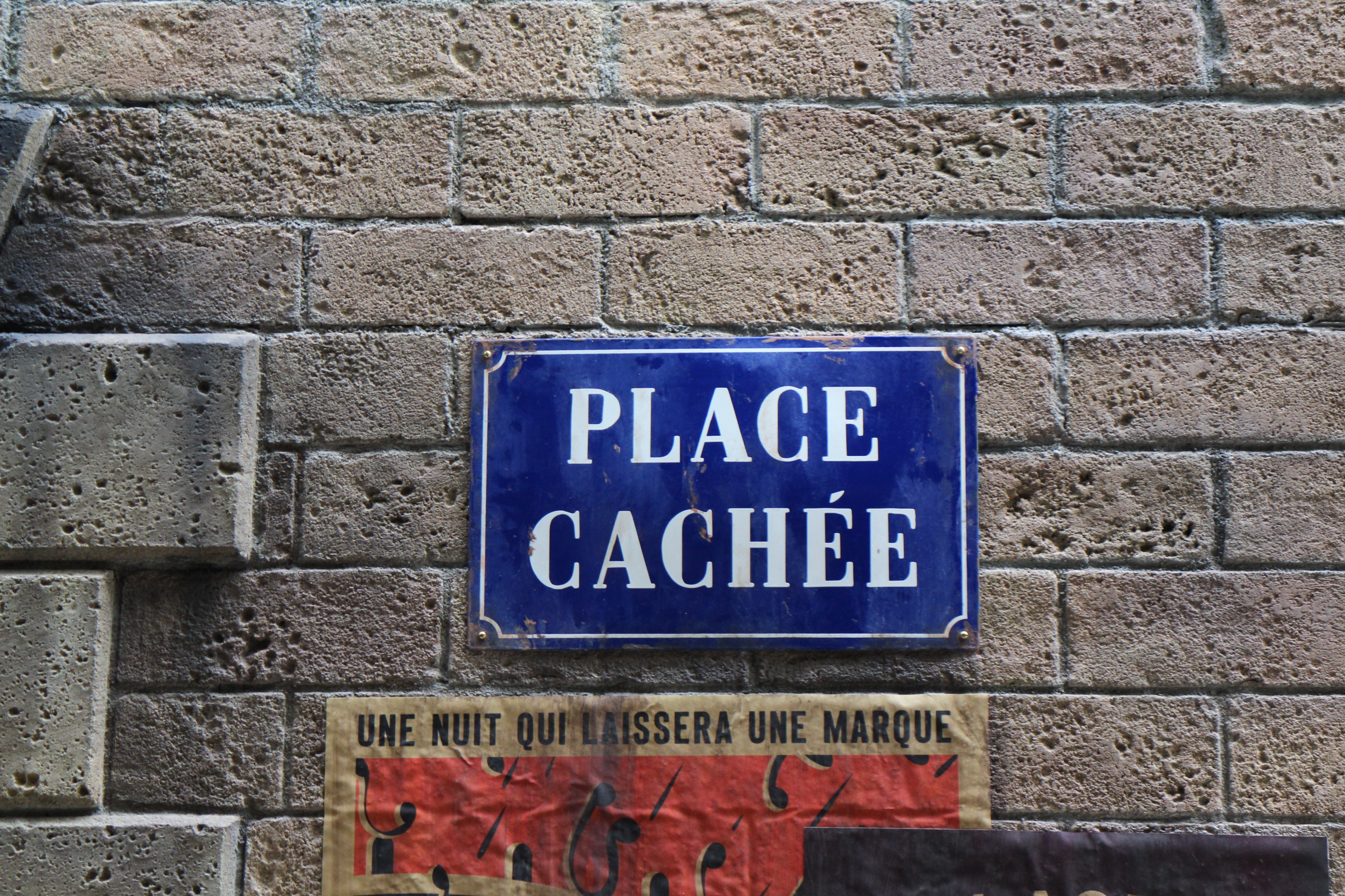 A blue Place Cachee sign with white writing seen on a brick wall in the exterior queue of the Ministry of Magic attraction in the Harry Potter area of Epic Universe.